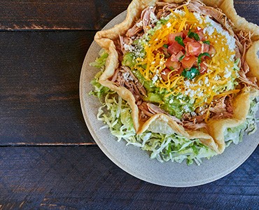 Tostada Bowl filled with Whole Beans, Meat, Guacamole, Sour Cream, Salsa Mexicana and Cheese